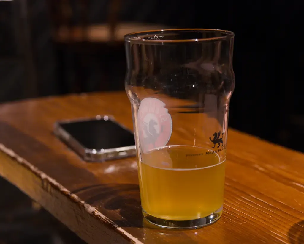 AI generated alt: A half-finished glass of pale beer on a varnished wooden bar top, a phone face-down beside it in the dark.